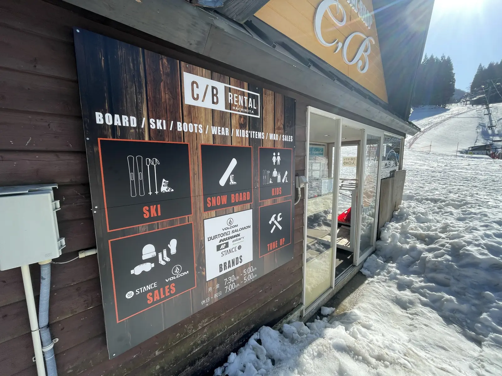 スキー場のゲレンデ前に建つレンタルショップの外壁。木目調の看板に、SKI、SNOW BOARD、KIDS、SALES、TUNE UPのアイコンと文字、営業時間が並ぶ。右奥にリフトとゲレンデが見える